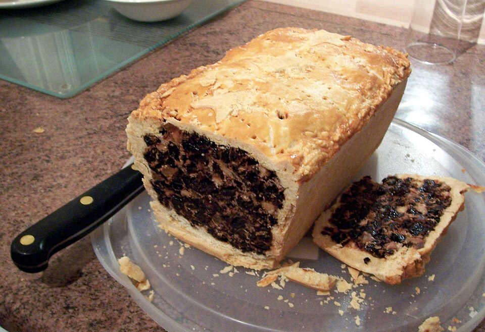 A black bun cut open to show the dense, dark fruit filling inside its pastry case